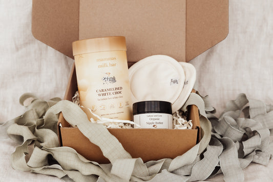 Box of skincare products including organic nipple butter, breastpads, and a white choc lactation powder on a soft fabric background.