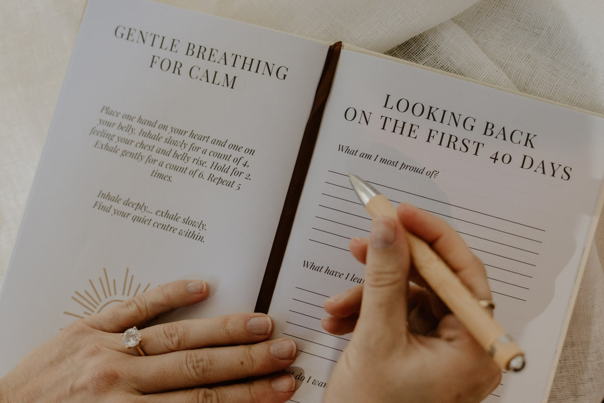 Postpartum journal open to gentle breathing and Looking Back on the First 40 Days prompts, with a pen in hand