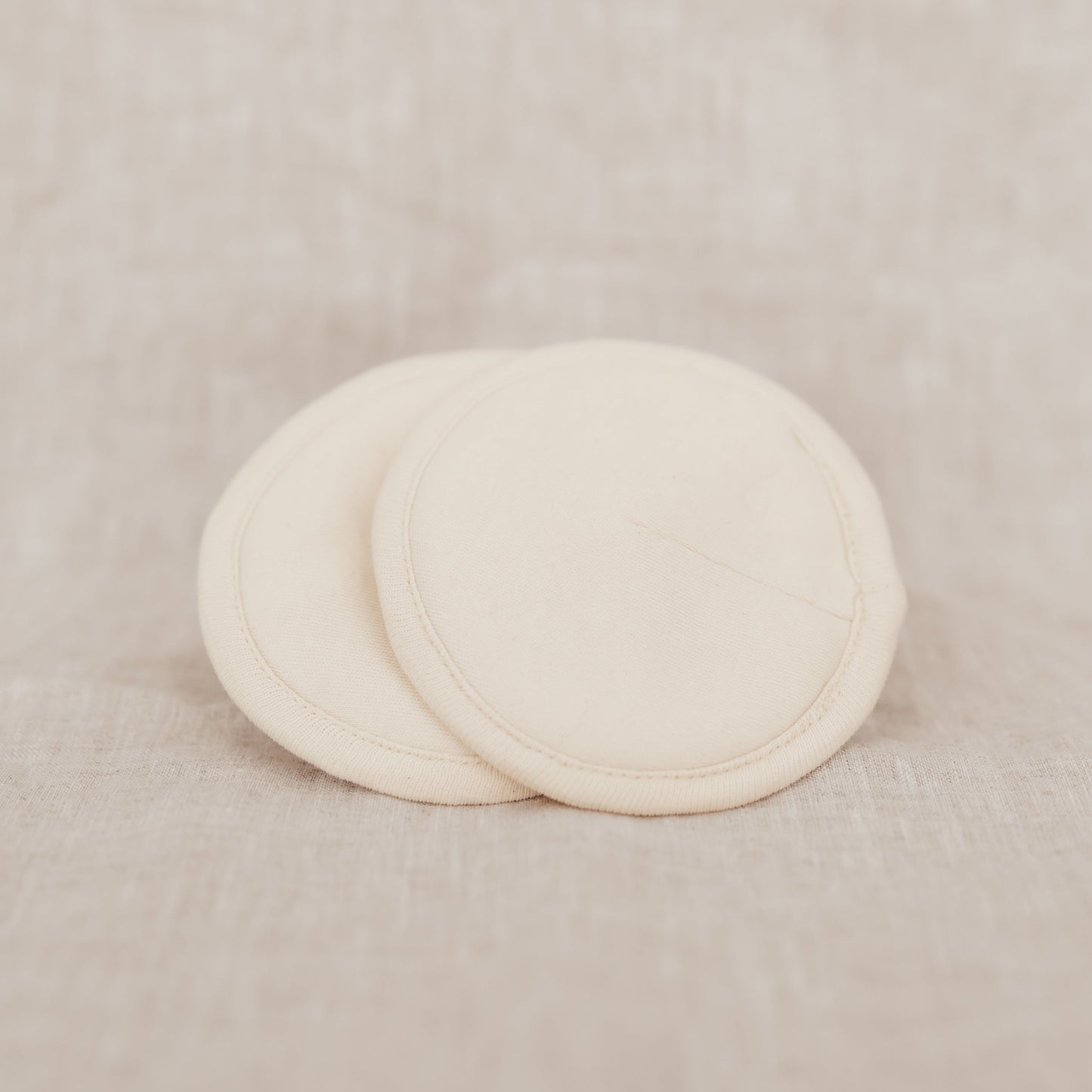 Two beige organic cotton breast pads on a linen background