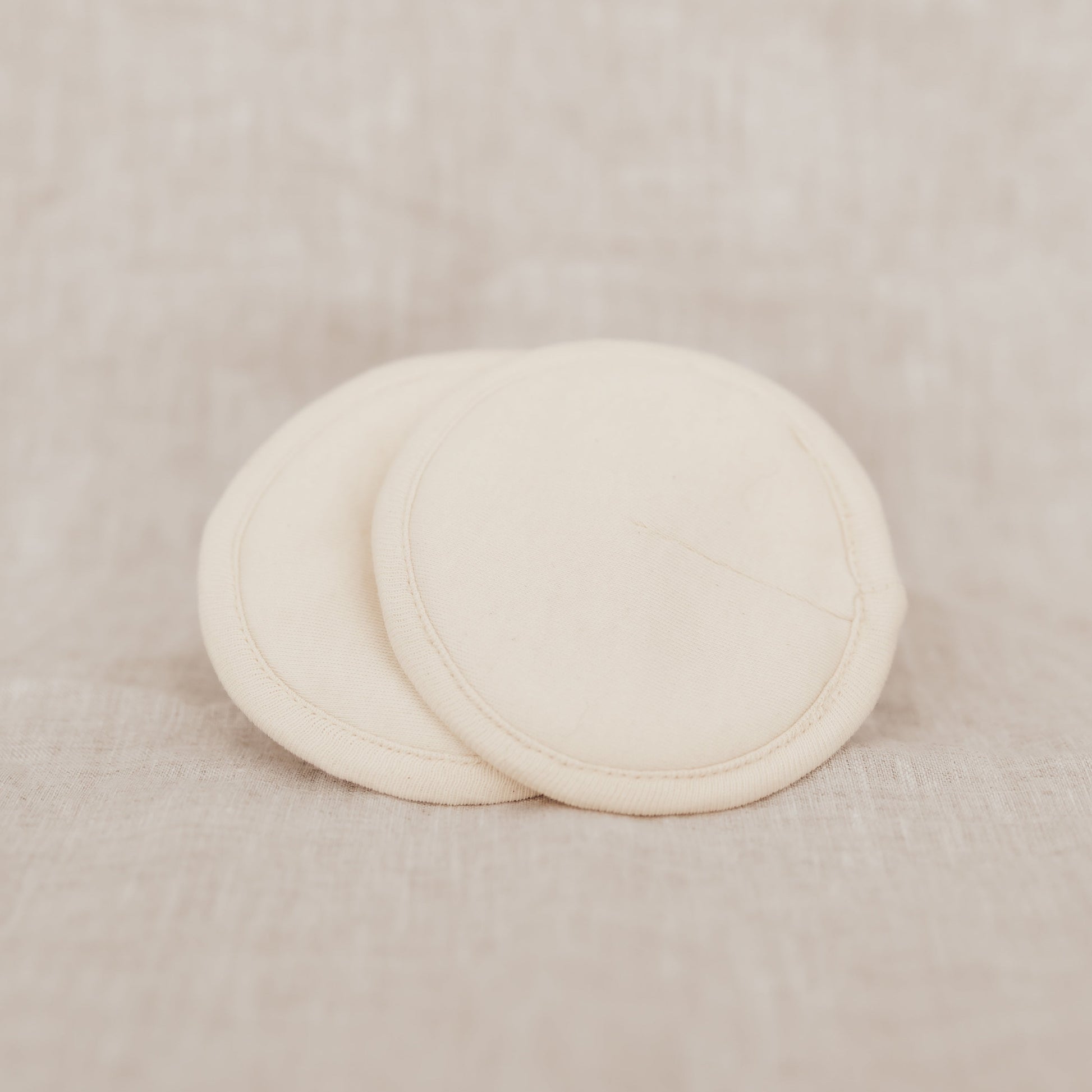 Two beige organic cotton breast pads on a linen background