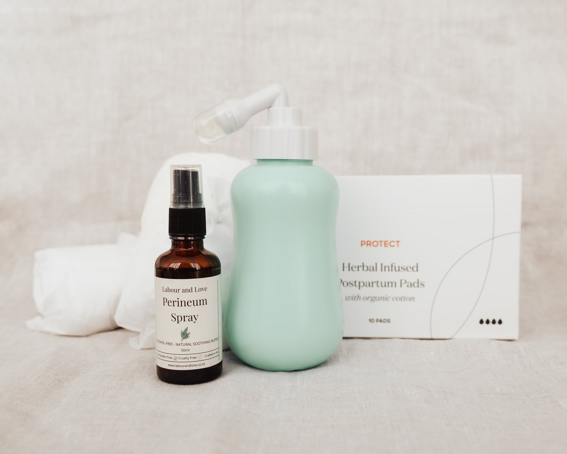 Four products including a peri bottle, perineum relief spray, organic cotton disposable postpartum undies and box of organic postpartum pads on a neutral background. 
