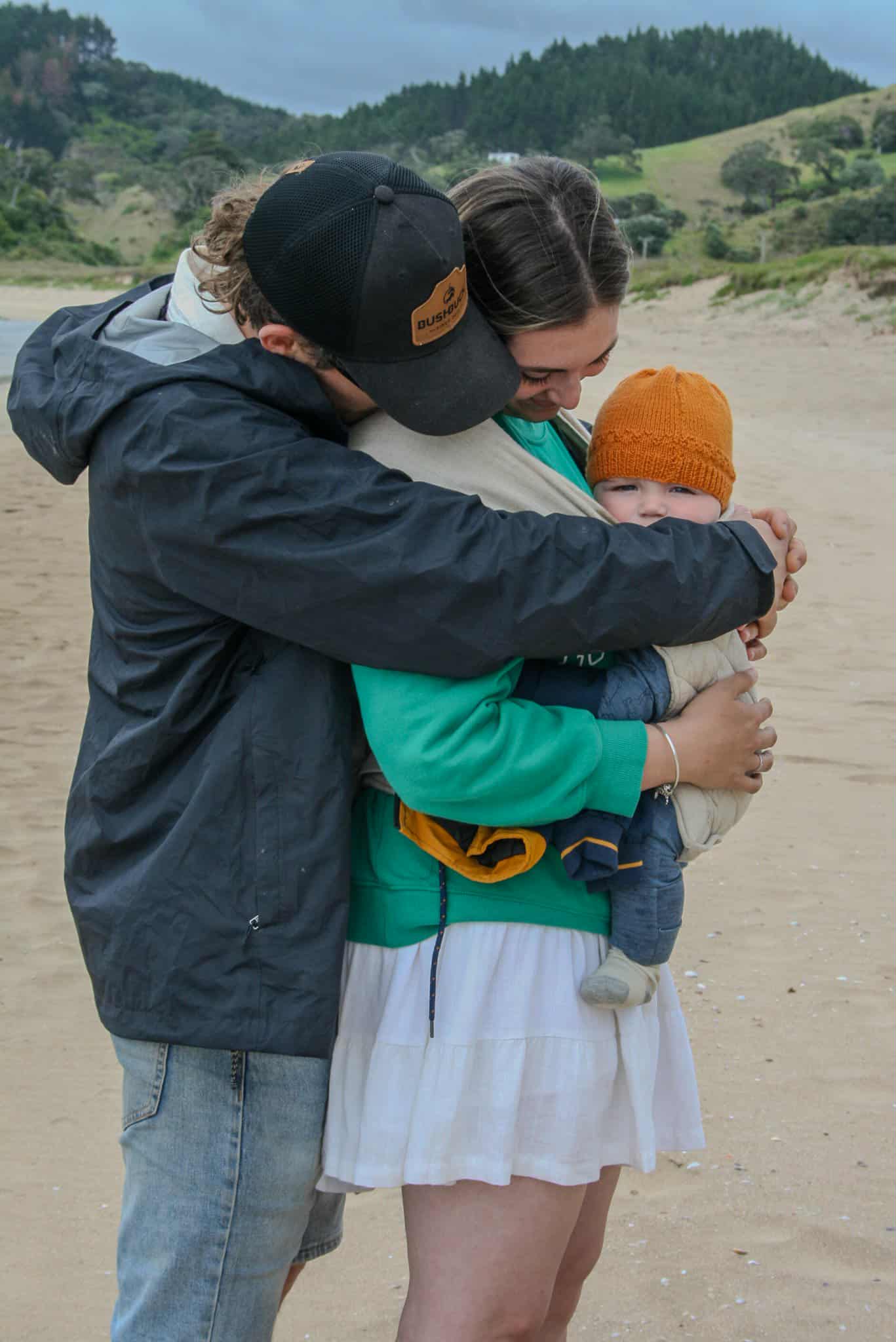 Two adults hugging a child on a sandy beach with green hills in the background