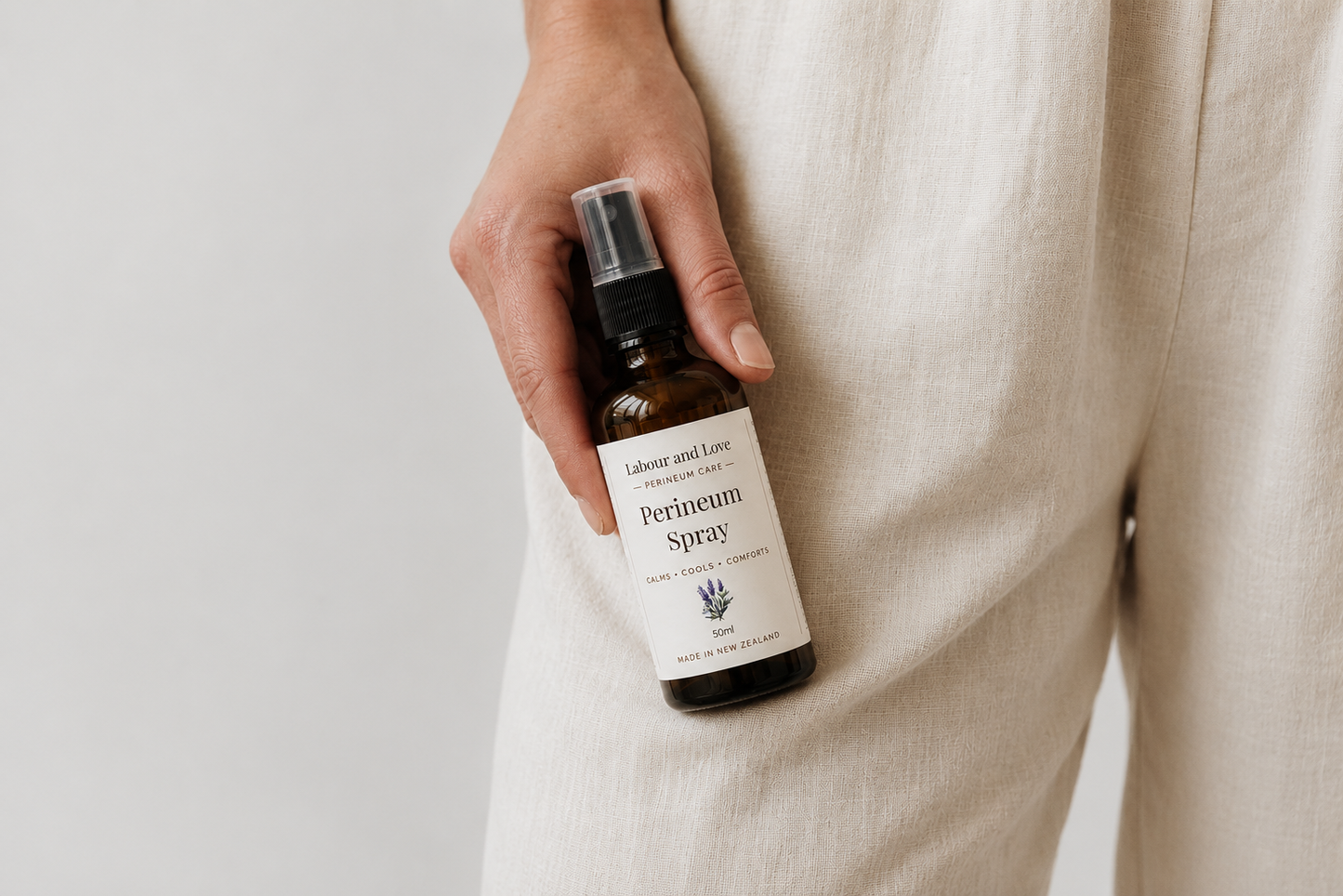 Hand holding an amber glass bottle of Labour & Love Perineum Relief Spray — gentle alcohol-free witch hazel & lavender hydrosol postpartum perineal spray, made in NZ